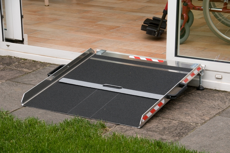 Eine tragbare, silberne Rollstuhlrampe aus Metall mit schwarzer Anti-Rutsch-Beschichtung überbrückt die Türschwelle zwischen einer Terrasse und dem Innenraum. Die Rampe verfügt über seitliche Schutzränder, rot-weiße Sicherheitsmarkierungen und praktische Tragegriffe an den Seiten. Im Hintergrund ist im Türrahmen ein Teil eines Rollstuhls zu erkennen, der die Rampe nutzt. Diese mobile Lösung ermöglicht einen barrierefreien Zugang über kleine Stufen oder Absätze hinweg.