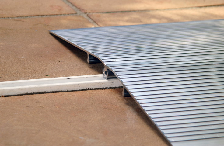 Eine ältere Dame im Rollstuhl wird von einer Begleitperson über eine breite, silberne Aluminiumrampe geschoben, die den Höhenunterschied zwischen dem gepflasterten Außenbereich und der gläsernen Eingangstür eines modernen Gebäudes überbrückt. Die flache Rampe mit rutschfester Oberfläche ermöglicht einen barrierefreien Zugang und verfügt über seitliche Begrenzungen für zusätzliche Sicherheit. Der glänzende Metallhandlauf an der Hauswand bietet zudem Halt beim Begehen des Eingangs.