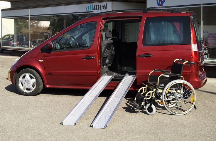Ein roter Van steht mit geöffneter Schiebetür auf einem Parkplatz, wobei zwei schmale Teleskoprampen aus Aluminium den Einstieg für einen Rollstuhl barrierefrei überbrücken. Neben den Rampen wartet ein leerer Rollstuhl auf dem Asphalt, der bereit ist, über die Schienen in das Fahrzeug geschoben zu werden. Diese mobile Verladehilfe ermöglicht einen flexiblen Transport von Rollstuhlnutzern ohne festen Fahrzeugumbau.