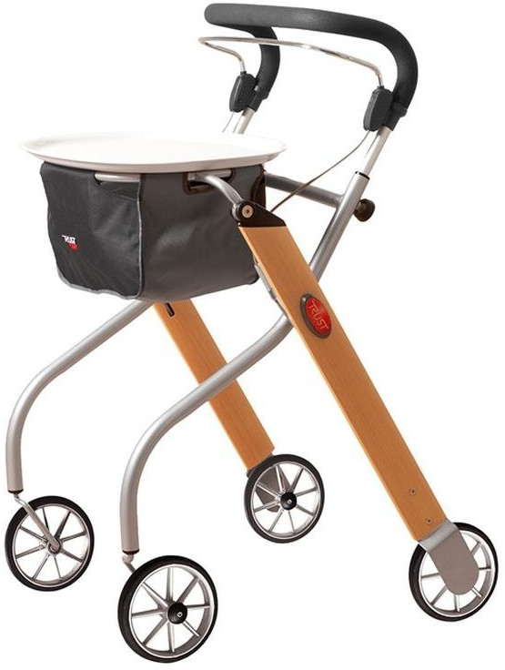 Ein eleganter Wohnraum-Rollator kombiniert einen silberfarbenen Aluminiumrahmen mit breiten Standbeinen in heller Holzoptik. Er ist mit vier großen, schmalen Speichenrädern und einem durchgehenden, schwarz gepolsterten Schiebegriff mit integriertem Bremsbügel ausgestattet. Auf der Oberseite befindet sich ein rundes, weißes Tablett, unter dem eine dunkelgraue Stofftasche für den Transport von Utensilien befestigt ist. Das Design ist speziell auf die sichere und stilvolle Nutzung innerhalb der Wohnung ausgerichtet.
