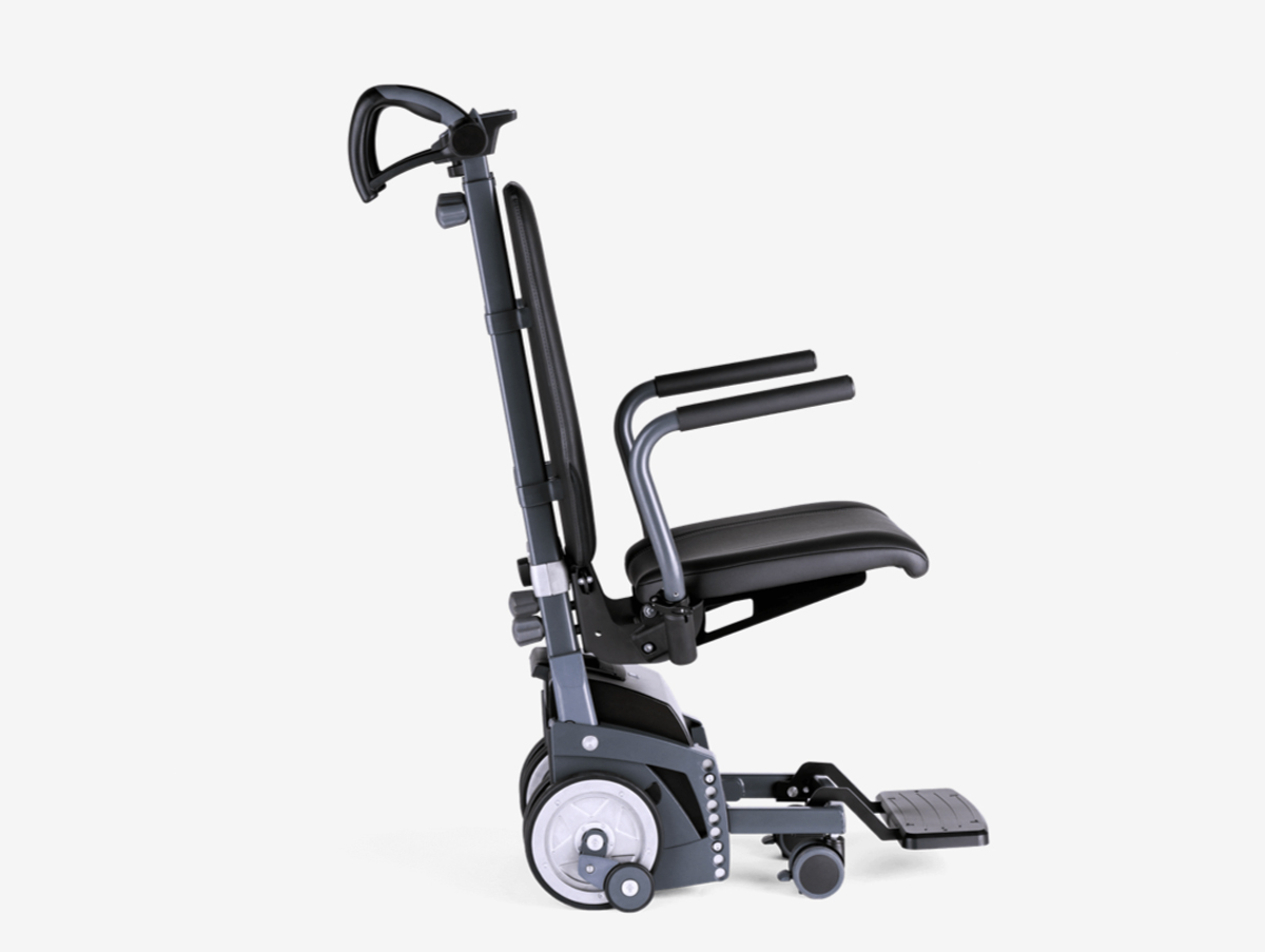 Das Bild zeigt die Nahaufnahme einer Person, die einen elektrischen Treppensteiger an den ergonomisch geformten Griffen führt. Zwischen den Griffen befindet sich eine Bedieneinheit mit einem digitalen Display zur Steuerung und Überwachung des Geräts. Die Szene verdeutlicht die Handhabung des Hilfsmittels während des Transports auf einer Treppe.