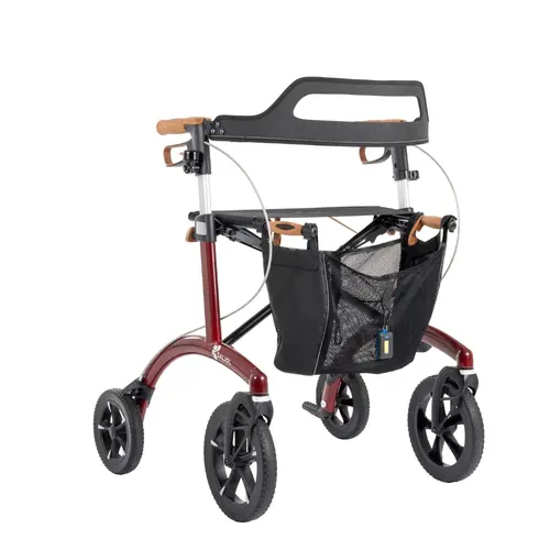 Ein moderner, vierrädriger Rollator in Dunkelgrün steht auf weißem Untergrund. Er besitzt braune Griffe, eine schwarze Sitzfläche mit schwarzem Rückengurt und eine vordere Einkaufstasche mit braunem Deckel. Oben rechts befindet sich eine dunkle, runde Grafik mit der Aufschrift „Jetzt neu! Inklusive Rückengurt, Stockhalter, LED-Licht, Tasche, Klingel“. Die Tasche hat ein angebrachtes LED-Licht und der Rollator ist mit Bremskabeln und sichtbaren Bremshebeln ausgestattet. Die Räder sind groß und schwarz.